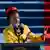 A young African American woman dressed in a yellow coat and wearing a red headband speaks into the microphone. It is American National Youth Poet Laureate, Amanda Gorman