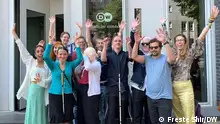 Zu sehen ist das Innoklusio Team mit einem Teil des DW Diversity Managements, wie sie vor dem Eingang des DW Gebäudes am Berliner Standort stehen. Sie halten die Arme hoch, winken und lächeln. 