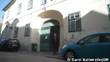 Der Physiker und Buchautor Marko Ivanovic steht vor dem Haus, das einst seiner Familie gehörte. Das grüne Tor hinter ihm steht halb offen. Zwei Autos sind am Rand der engen Straße geparkt. 
